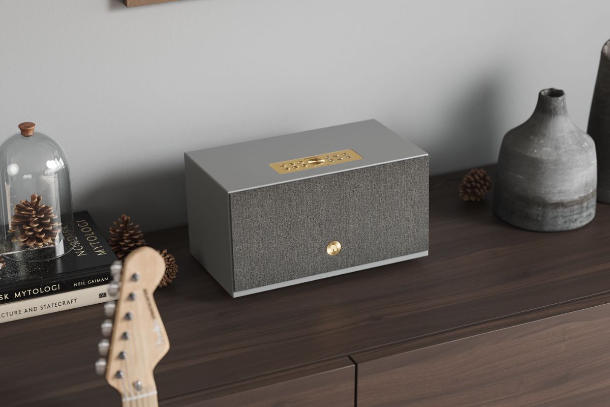 Kompakter HiFi-Lautsprecher mit Stofffront und goldfarbenen Bedienelementen steht auf einem Sideboard neben Büchern, Dekoration und einer Gitarre in einem modern eingerichteten Wohnraum.