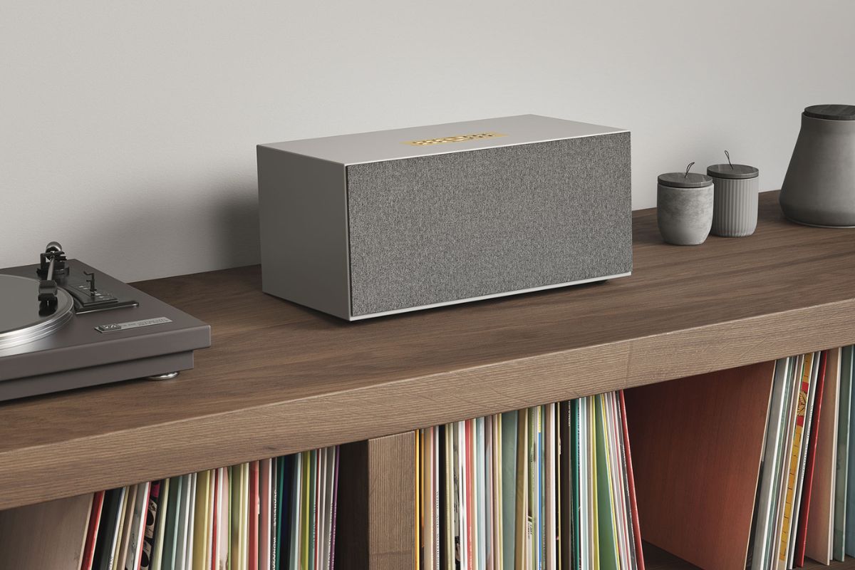 Moderner HiFi-Lautsprecher mit grauer Stofffront steht auf einem Holz-Sideboard neben einem Plattenspieler, dekorativen Gefäßen und einer Schallplattensammlung in einem wohnlichen Musikzimmer