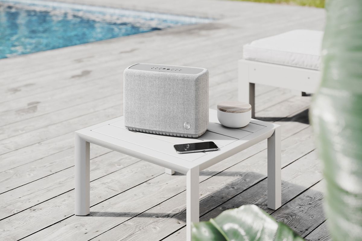 Tragbarer Multiroom-Lautsprecher in Hellgrau von Audio Pro auf einem weißen Terrassentisch am Pool